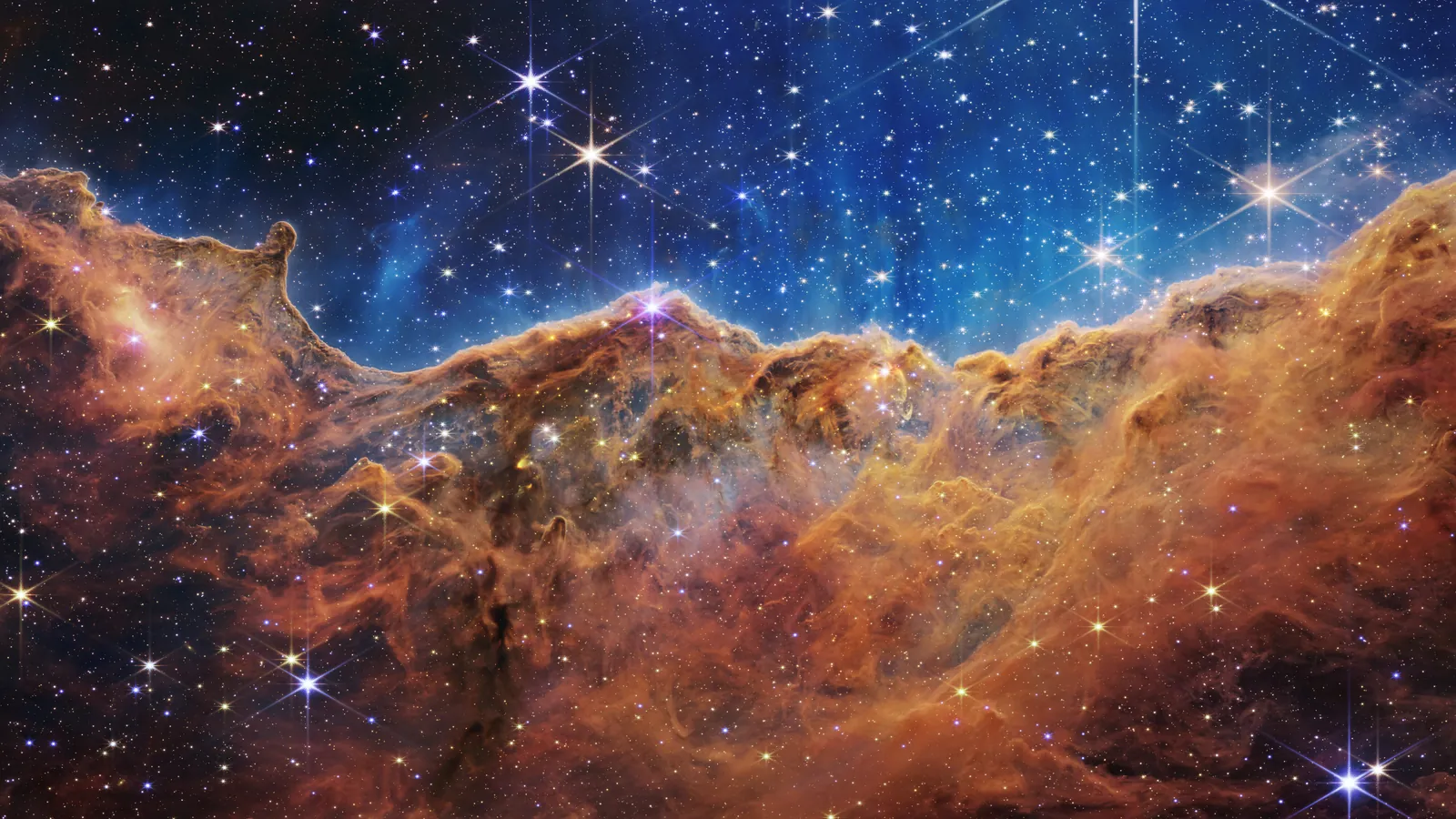 Webb's view of the Cosmic Cliffs in the Carina Nebula, an orange-brown ridge of gas and dust beneath a blue, star-filled sky