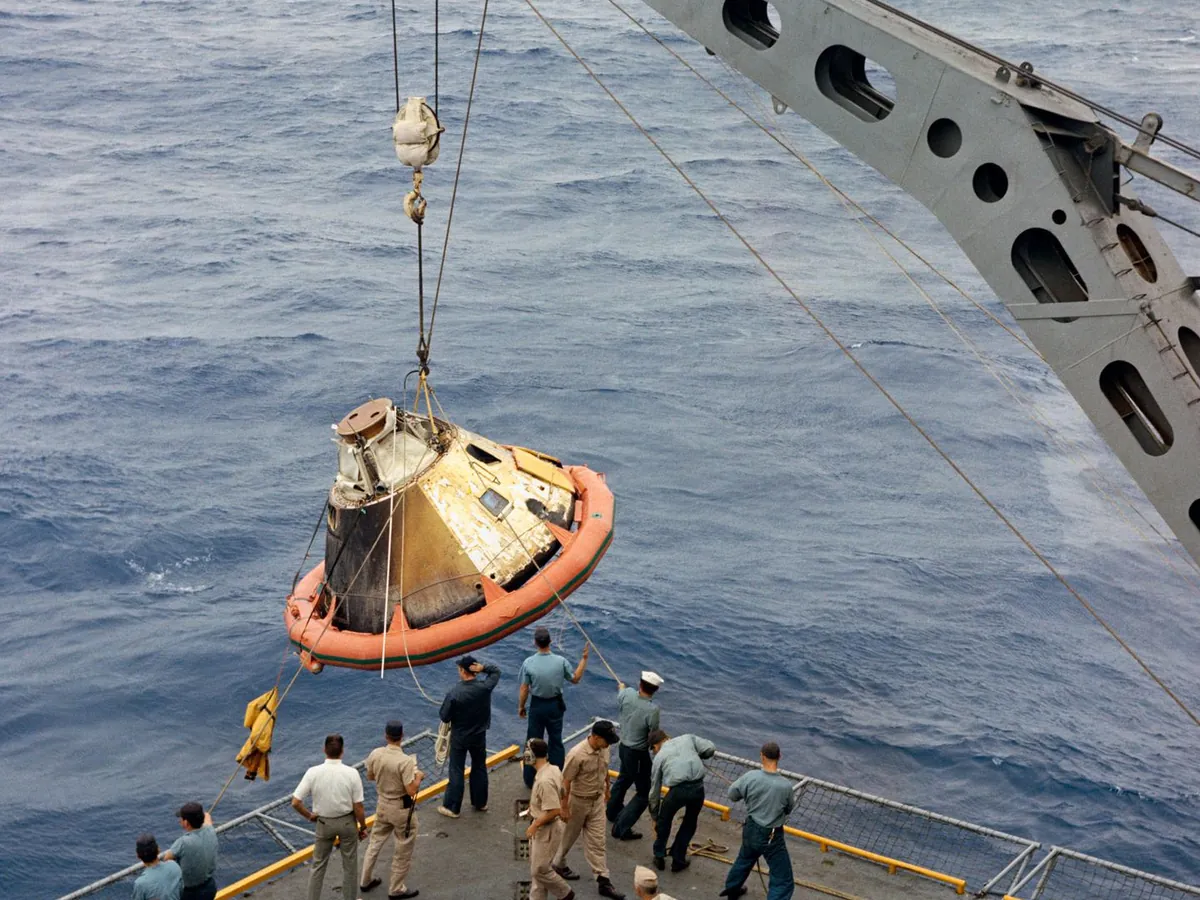 The Apollo 4 command module, ringed by an orange flotation collar, being hoisted by crane onto the USS Bennington, its heat shield visibly charred