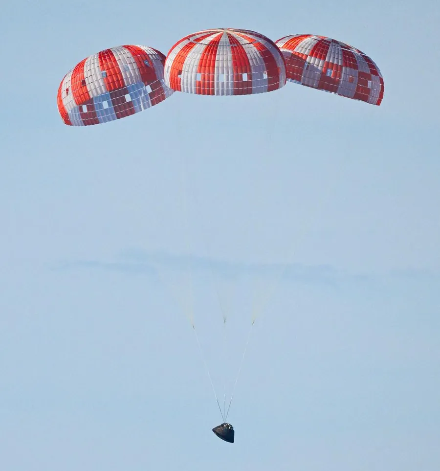 The Orion spacecraft descending under three red-and-white main parachutes against a pale blue sky