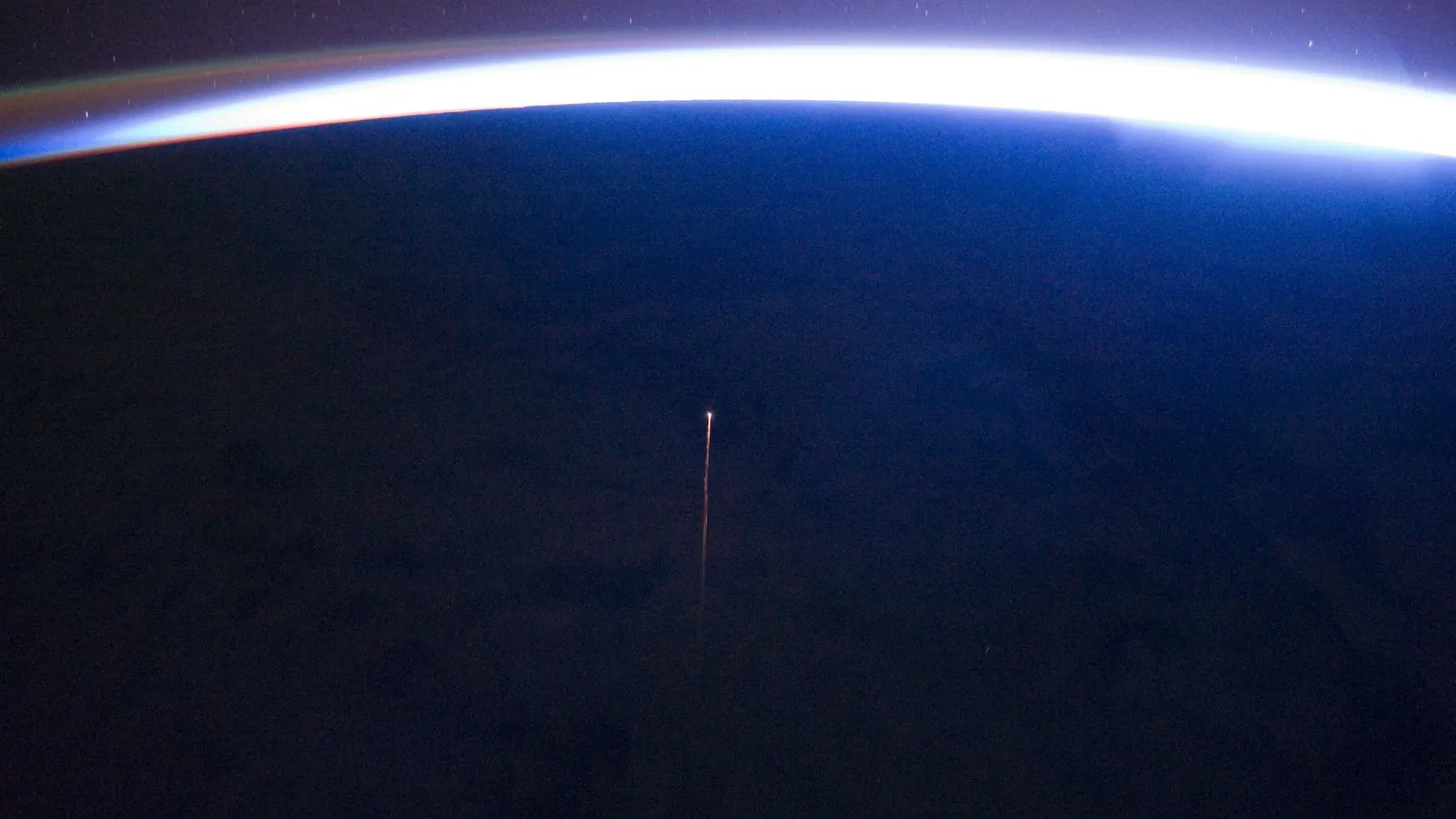 Earth's night side seen from the International Space Station, a bright band of airglow along the horizon and a thin orange plasma trail from a Progress cargo ship reentering below